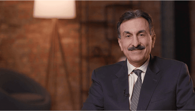 Man in black suit with black and grey patterned tie who is smiling