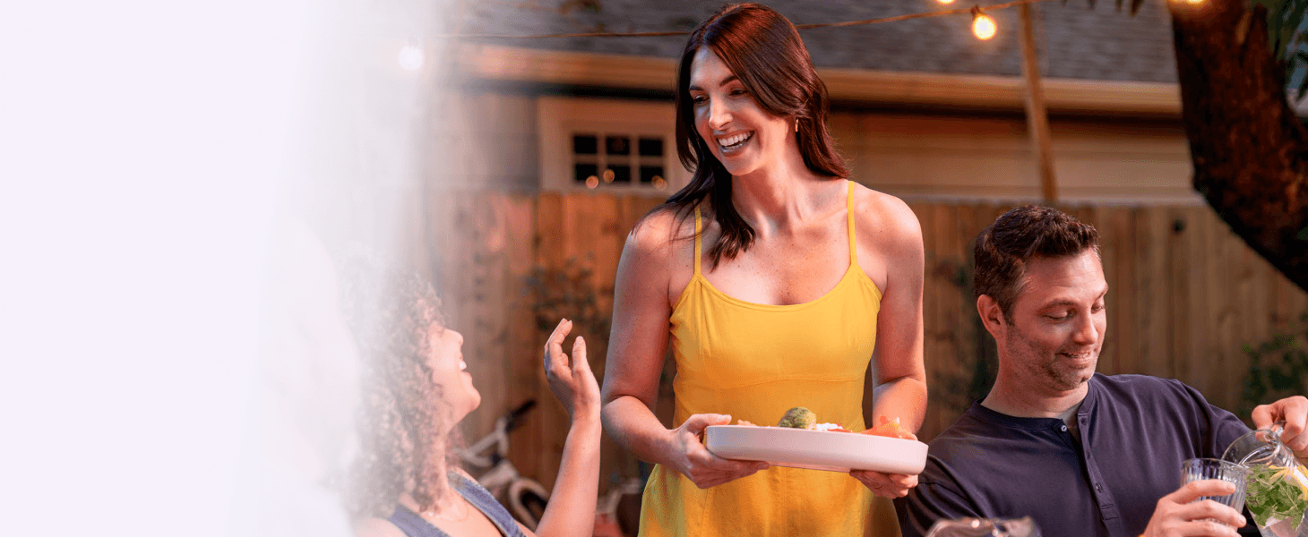 A woman carrying a dish of food while socializing with friends at a backyard dinner gathering