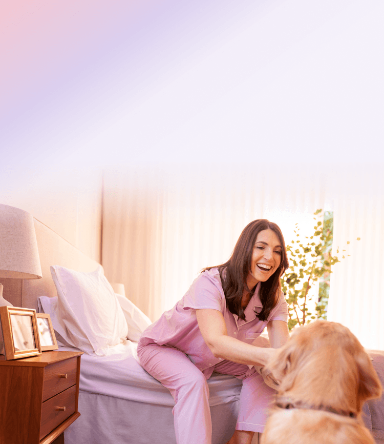 A woman in pajamas sitting on her bed laughing while petting her dog