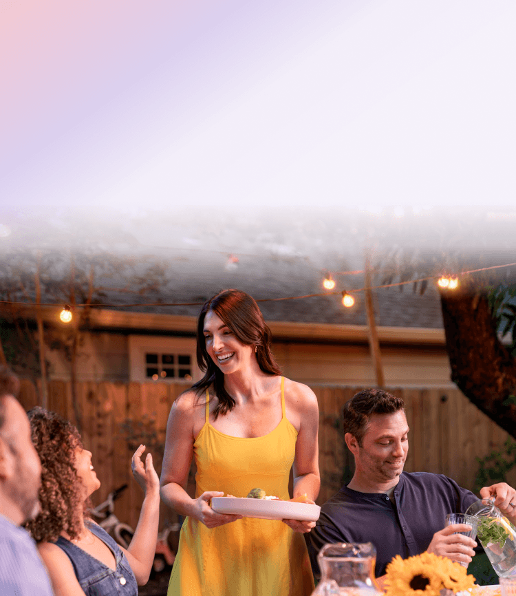 A woman carrying a dish of food while socializing with friends at a backyard dinner gathering